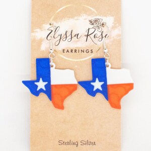 Puede incluir: Un par de pendientes con la forma del estado de Texas, con el diseño de la bandera de Texas. Los pendientes son azules, blancos y naranjas con una estrella blanca. Cuelgan de ganchos plateados. El fondo incluye el texto "Elyssa Rose Earrings" y "Sterling Silver".
