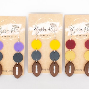 May include: Three pairs of earrings with a football theme. Each earring has a football shape at the bottom, a dark grey circle, and a coloured circle at the top. The colours are purple, yellow, and maroon.