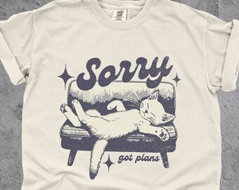 May include: A white t-shirt with a vintage-style illustration of a cat sleeping on a couch. The text "Sorry got plans" is printed below the cat.