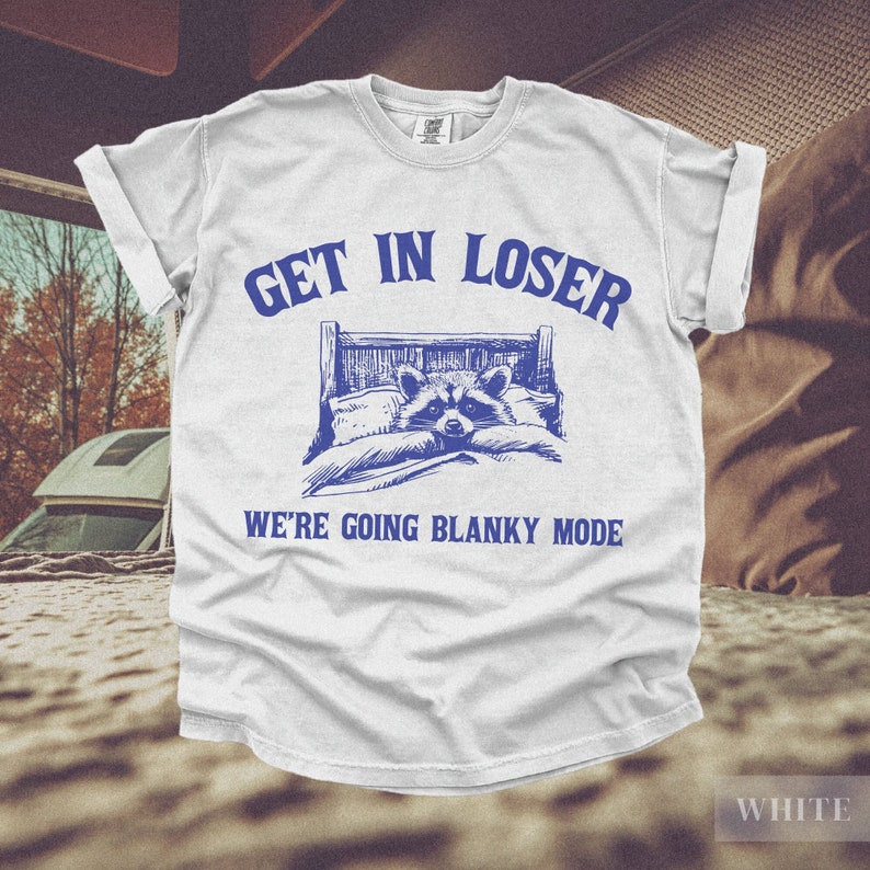 K&ouml;nnte beinhalten: Wei&szlig;es T-Shirt mit blauem Text, der "Get in loser, we're going blanky mode" lautet. Ein Cartoon-Waschb&auml;r liegt in einem Bett mit einer Decke.