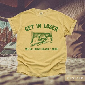 K&ouml;nnte beinhalten: Ein gelbes T-Shirt mit gr&uuml;nem Text, der "Get in loser, we're going blanky mode" lautet. Der Text befindet sich &uuml;ber einem Cartoon-Bild eines Waschb&auml;ren im Bett.