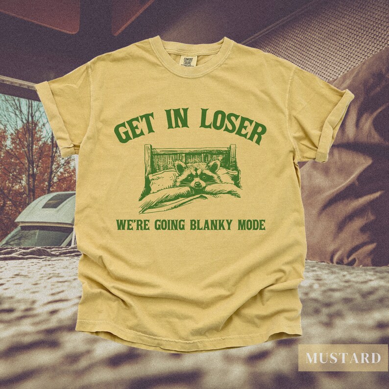 K&ouml;nnte beinhalten: Ein gelbes T-Shirt mit gr&uuml;nem Text, der "Get in loser, we're going blanky mode" lautet. Der Text befindet sich &uuml;ber einem Cartoon-Bild eines Waschb&auml;ren im Bett.
