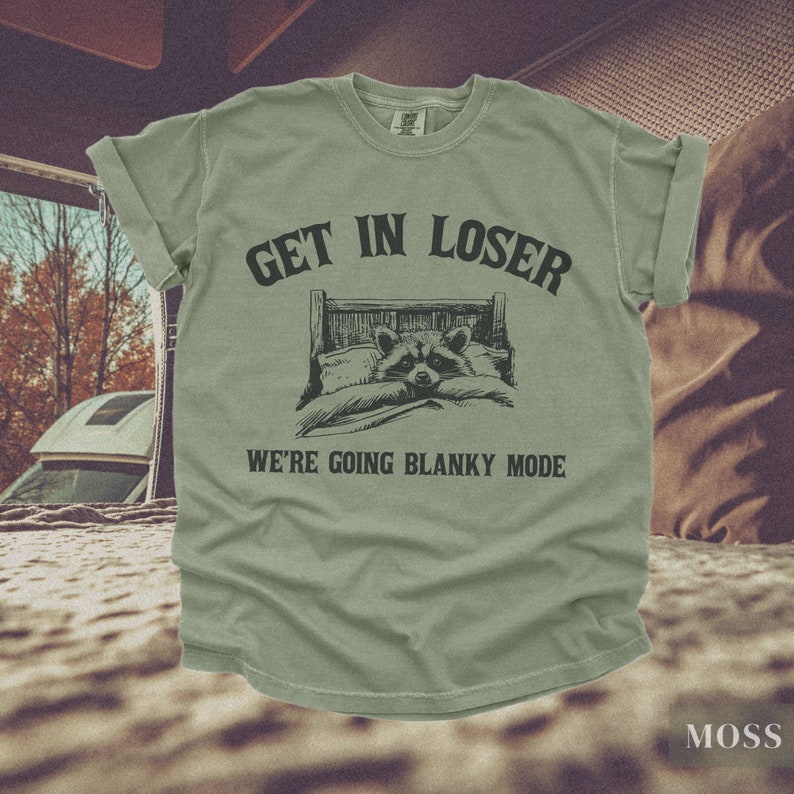 K&ouml;nnte beinhalten: Ein gr&uuml;nes T-Shirt mit dem Text "Get in loser, we're going blanky mode" und einem Cartoon-Waschb&auml;ren im Bett.