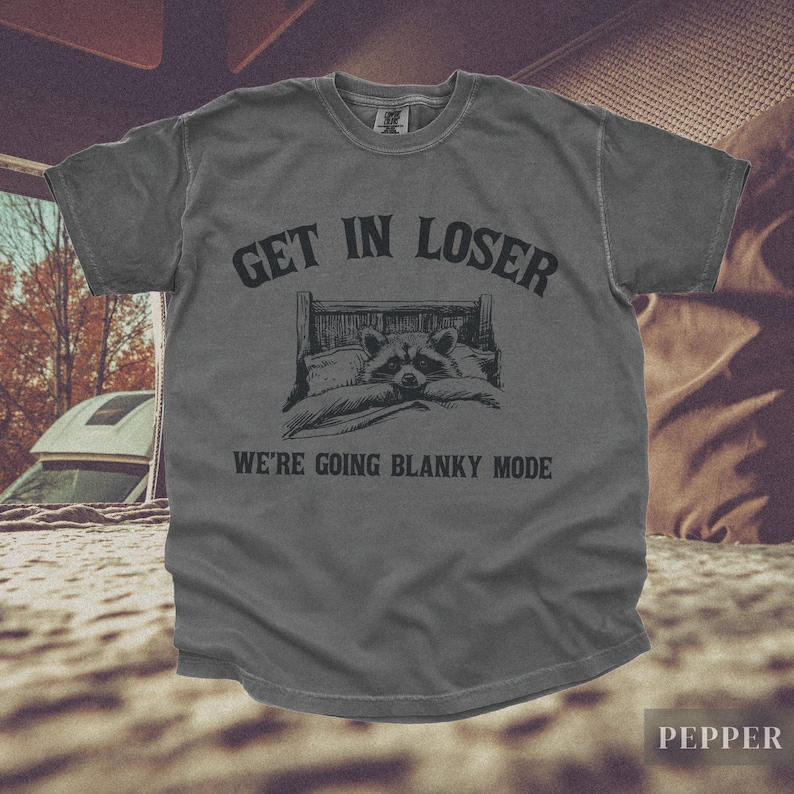 K&ouml;nnte beinhalten: Ein dunkelgraues T-Shirt mit dem Text "Get in loser, we're going blanky mode" in Schwarz. Ein Cartoon-Waschb&auml;r liegt in einem Bett mit einer Decke.