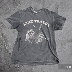 May include: A dark gray t-shirt with a white graphic of a raccoon with the text "STAY TRASHY" above it.