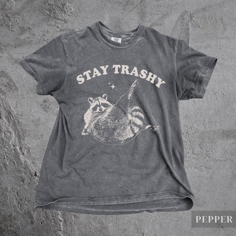 May include: A dark gray t-shirt with a white graphic of a raccoon with the text "STAY TRASHY" above it.