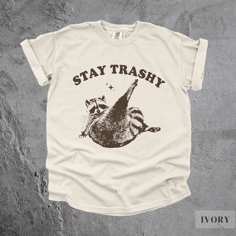 May include: A cream colored t-shirt with a brown raccoon graphic and the text "STAY TRASHY" in brown letters. The t-shirt is made of a soft, comfortable material.