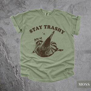 May include: A green t-shirt with a brown raccoon graphic and the text "STAY TRASHY" in brown letters.