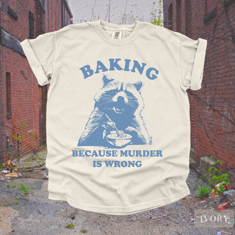 May include: A light beige t-shirt with a blue raccoon graphic and the text "Baking because murder is wrong".