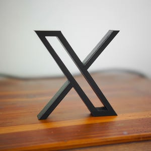 Puede incluir: Una escultura geométrica negra en forma de X. La escultura está hecha de material sólido y se encuentra sobre una superficie de madera. El fondo es blanco borroso.