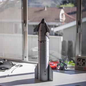 May include: A silver and black model of a spacecraft with a pointed nose and four fins. The model is sitting on a white surface.