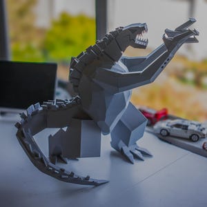 May include: A gray, geometric Godzilla sculpture with an open mouth and raised arms. The model is on a white surface, with toy cars in the background. The sculpture has a detailed, angular design.