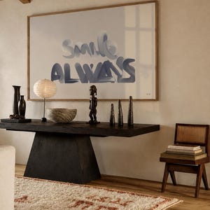 Può includere: Una grande opera d'arte incorniciata con il testo "SMILE ALWAYS" in un carattere blu stilizzato. L'opera è esposta sopra una console in legno scuro che sostiene una lampada di carta rotonda, una ciotola testurizzata e sculture decorative.