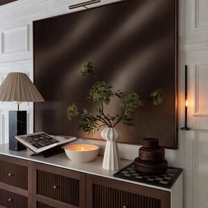 Può includere: Scena d'interni moderna con un grande specchio con cornice marrone, un vaso bianco con verde e una candela accesa in una ciotola bianca. Un mobile in legno scuro con pomelli dorati e una base per lampada in marmo.