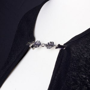 May include: A silver-toned shawl clip with a leaf design. The clip is attached to a black garment, showcasing the accessory's decorative function. The clip has two end pieces that attach to the garment and a chain of leaves in the center.
