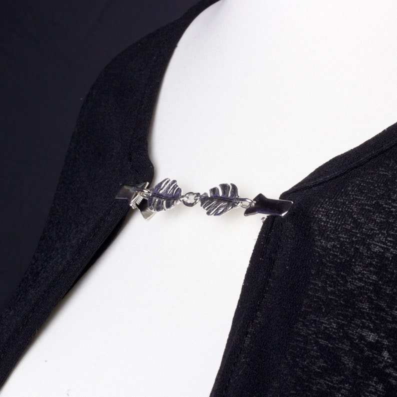 May include: A silver-toned shawl clip with a leaf design. The clip is attached to a black garment, showcasing the accessory's decorative function. The clip has two end pieces that attach to the garment and a chain of leaves in the center.