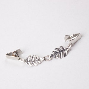 May include: A silver-toned shirt clip featuring three decorative leaf charms. The clip has two metal clasps connected by a chain with the leaf accents. The leaves have detailed veining, adding a naturalistic touch to the accessory.