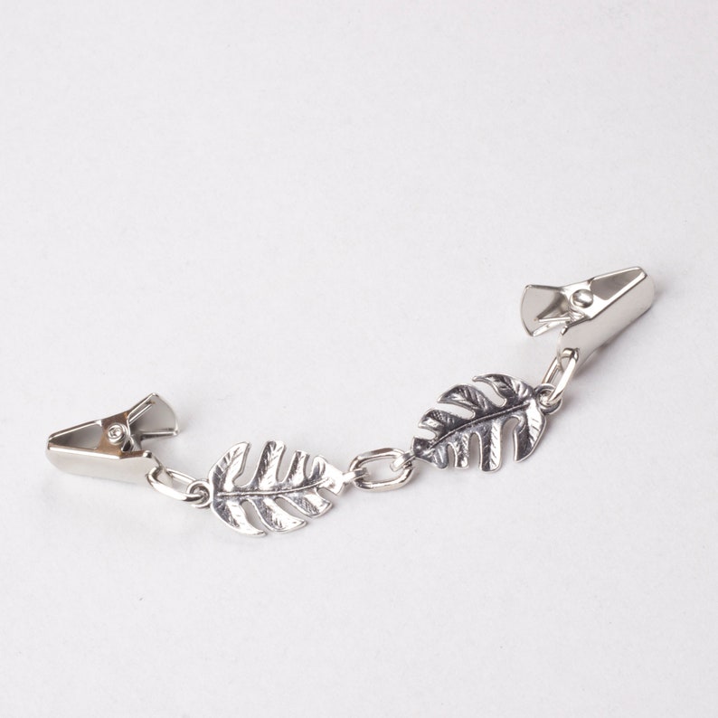 May include: A silver-toned shirt clip featuring three decorative leaf charms. The clip has two metal clasps connected by a chain with the leaf accents. The leaves have detailed veining, adding a naturalistic touch to the accessory.