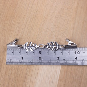 May include: Silver-toned leaf-shaped shirt clips with chain links and two metal clips. The clips are approximately 4 inches long, as measured by the ruler in the image. The design features detailed leaf patterns.
