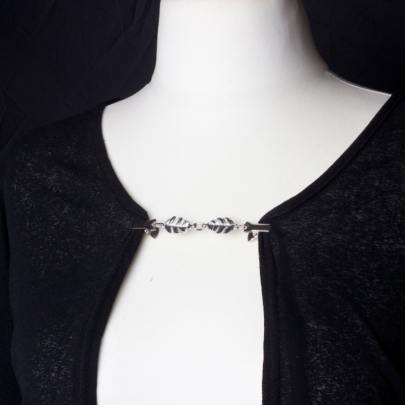 May include: A silver-toned cardigan clip with a leaf design, attached to a black cardigan. The clip is made of metal and features a series of leaf-shaped charms connected by small chains. The cardigan is displayed on a white mannequin.