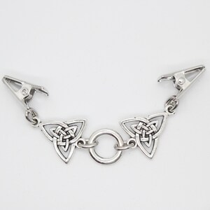 Puede incluir: Pinza de ropa de metal plateado con dise&ntilde;o de nudo celta. La pinza presenta dos colgantes triangulares de nudo celta conectados por un peque&ntilde;o anillo. Cada extremo tiene una pinza met&aacute;lica para sujetar. El fondo es blanco liso.