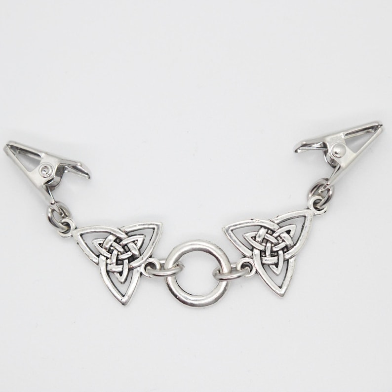 Puede incluir: Pinza de ropa de metal plateado con dise&ntilde;o de nudo celta. La pinza presenta dos colgantes triangulares de nudo celta conectados por un peque&ntilde;o anillo. Cada extremo tiene una pinza met&aacute;lica para sujetar. El fondo es blanco liso.