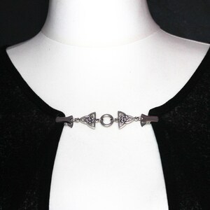 Puede incluir: Un broche plateado para capa con dise&ntilde;o de nudo celta. El broche est&aacute; compuesto por dos piezas decorativas en los extremos conectadas por una cadena con un anillo central. El broche est&aacute; sujeto a una prenda negra sobre un maniqu&iacute; blanco.