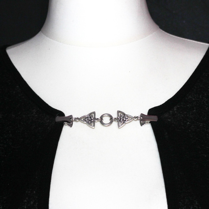 Puede incluir: Un broche plateado para capa con dise&ntilde;o de nudo celta. El broche est&aacute; compuesto por dos piezas decorativas en los extremos conectadas por una cadena con un anillo central. El broche est&aacute; sujeto a una prenda negra sobre un maniqu&iacute; blanco.