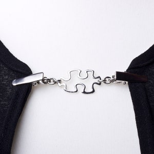 Könnte beinhalten: Ein silberfarbener Puzzle-Verschluss mit einer Kette, die zwei Clips verbindet. Der Verschluss ist so konzipiert, dass er zwei Stoffstücke zusammenhält.