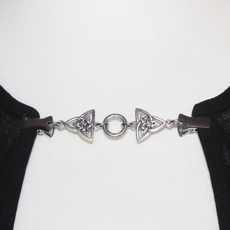 Puede incluir: Cierre de capa de metal plateado con dise&ntilde;o de nudo celta. El cierre presenta dos piezas triangulares con el dise&ntilde;o del nudo, conectadas por un anillo. El cierre est&aacute; sujeto a clips de cuero marr&oacute;n.