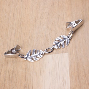 May include: A silver-colored metal clip with two alligator clips connected by a chain of three leaf-shaped charms. The clips are designed to hold items together. The background is a light brown wooden surface.