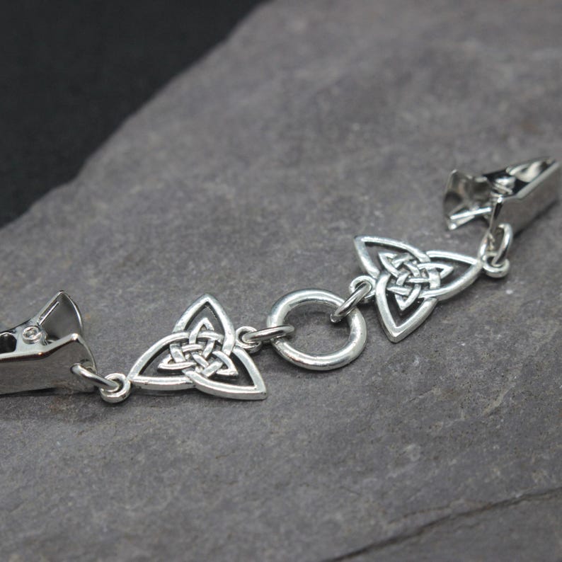 Puede incluir: Una pulsera de metal plateado con un dise&ntilde;o de nudo celta. La pulsera presenta tres dijes triangulares de nudo celta y dos cierres de clip. El dise&ntilde;o es sim&eacute;trico y el metal tiene un acabado pulido.