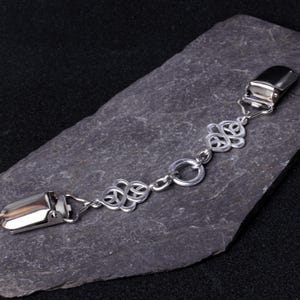Könnte beinhalten: Ein silberfarbener Strumpfhalter-Clip mit keltischem Knotenmuster. Der Clip hat zwei Metallverschlüsse, die durch eine Kette mit dekorativen Knoten und einem zentralen Ring verbunden sind. Der Strumpfhalter-Clip wird auf einer dunkelgrauen Steinoberfläche gezeigt.