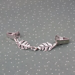 May include: A silver-toned metal clip-on accessory featuring two decorative leaf charms. The accessory has two clips on either end, connected by a chain with two leaf-shaped charms. The leaves have detailed vein patterns.
