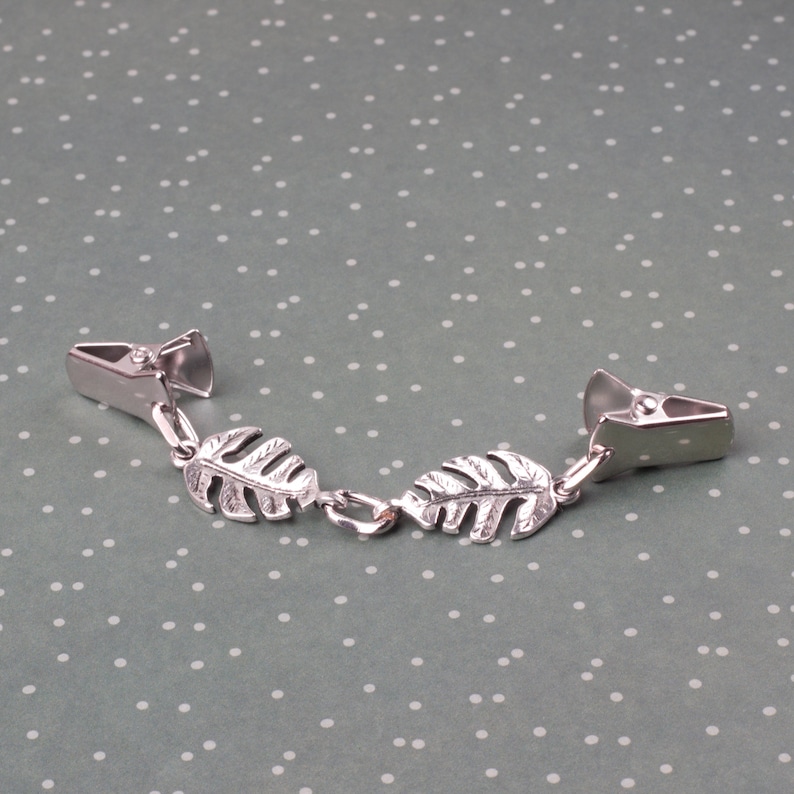 May include: A silver-toned metal clip-on accessory featuring two decorative leaf charms. The accessory has two clips on either end, connected by a chain with two leaf-shaped charms. The leaves have detailed vein patterns.