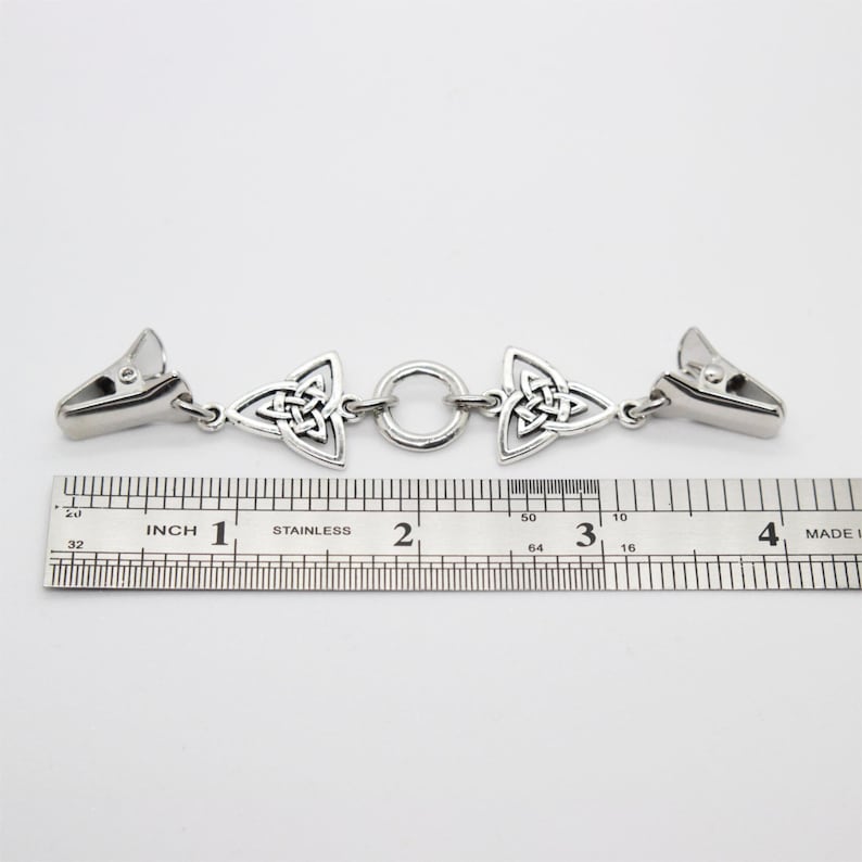 Puede incluir: Un clip de chal de nudo celta de metal plateado. El clip tiene dos extremos con un cierre y un dise&ntilde;o de nudo celta, conectados por un anillo. Mide aproximadamente 10 cm de largo.