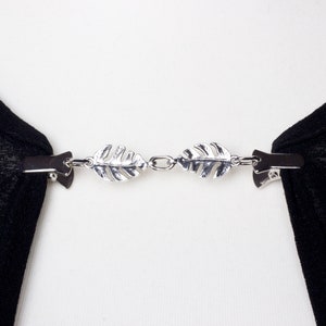 May include: A silver-toned scarf clip with a leaf design. The clip has two leaf-shaped ornaments connected by a chain. Each leaf is detailed with veining and attached to a clip on each end. The clip is holding black fabric.