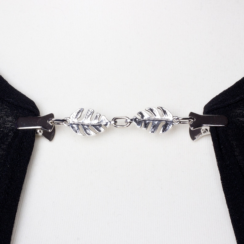 May include: A silver-toned scarf clip with a leaf design. The clip has two leaf-shaped ornaments connected by a chain. Each leaf is detailed with veining and attached to a clip on each end. The clip is holding black fabric.