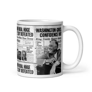 May include: White ceramic mug with a black and white newspaper print. Headlines read "NEW DEAL HUGE 130 DEFEATED" and "WASHINGTON GIVES CONFIDENCE VOT." The mug also features a black and white photograph of a man.