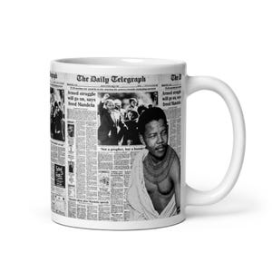 Könnte beinhalten: Eine weiße Keramiktasse mit einem Schwarzweißfoto von Nelson Mandela auf der Vorderseite. Das Foto stammt aus einem Zeitungsartikel der Daily Telegraph. Die Schlagzeile lautet "Bewaffneter Kampf wird weitergehen, sagt freigelassener Mandela".