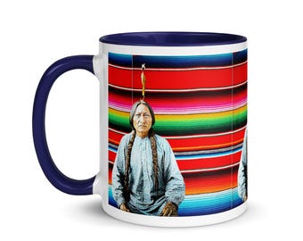 Może przedstawiać: Biały ceramiczny kubek z granatowym uchwytem i wnętrzem. Kubek przedstawia portret Indianina na tle kolorowego, pasiastym koca. Kubek ma standardowy rozmiar do gorących napojów.