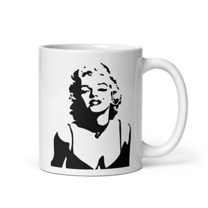 Puede incluir: Taza de cerámica blanca con una silueta en blanco y negro de una mujer. El diseño es un retrato estilizado, que muestra el rostro y la parte superior del cuerpo de la mujer. La taza tiene un asa curva y un acabado brillante.