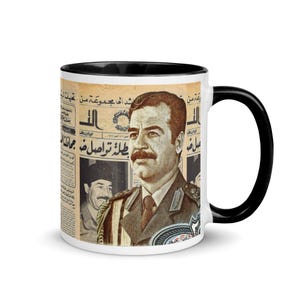 Può includere: Una tazza in ceramica bianca con bordo e manico neri. La tazza presenta una stampa di giornale vintage con un ritratto di Saddam Hussein. Il titolo del giornale recita "طلائع ثورة" in arabo.
