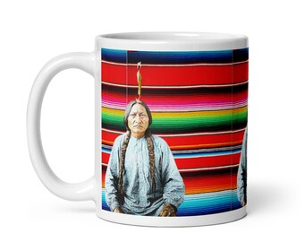 Może przedstawiać: Biały ceramiczny kubek do kawy z portretem Indianina z długimi warkoczami, na tle żywego, pasiastym kocu. Projekt kubka zawiera kolorowe tło z czerwonych, zielonych i żółtych pasków.