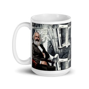 May include: White ceramic mug featuring a portrait of Karl Marx in a black suit and a long white beard. The background is a black and white illustration. The mug has a white handle.
