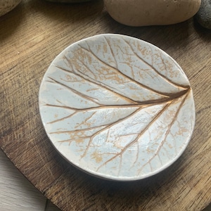 May include: A white ceramic dish shaped like a leaf with gold leaf veins. The dish is on a wooden surface.