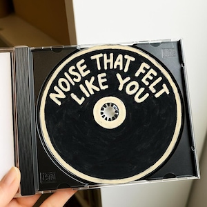 Puede incluir: Un CD negro con las palabras "NOISE THAT LIKE YOU FELT" en letras blancas. El CD está en una funda de plástico transparente, sostenida por una mano. El CD tiene un centro blanco y una superficie negra.