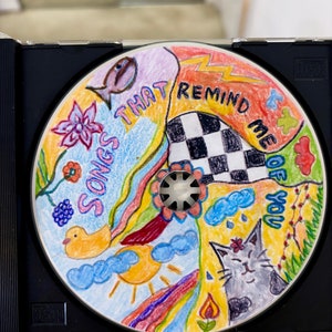 May include: A colorful CD with hand-drawn illustrations of a fish, a flower, a duck, a sun, a rainbow, a cat, a checkered pattern, and clouds. The text on the CD reads "Songs That Remind Me Of You".