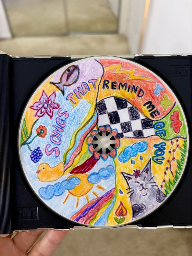 May include: A colorful CD with hand-drawn illustrations of a fish, a flower, a duck, a sun, a rainbow, a cat, a checkered pattern, and clouds. The text on the CD reads "Songs That Remind Me Of You".