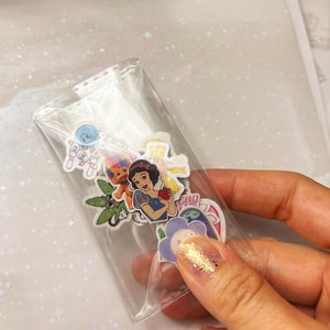 May include: A clear plastic bag filled with colorful stickers featuring cartoon characters like Snow White, a troll, and a flower. The stickers are on a white background with a glittery texture.